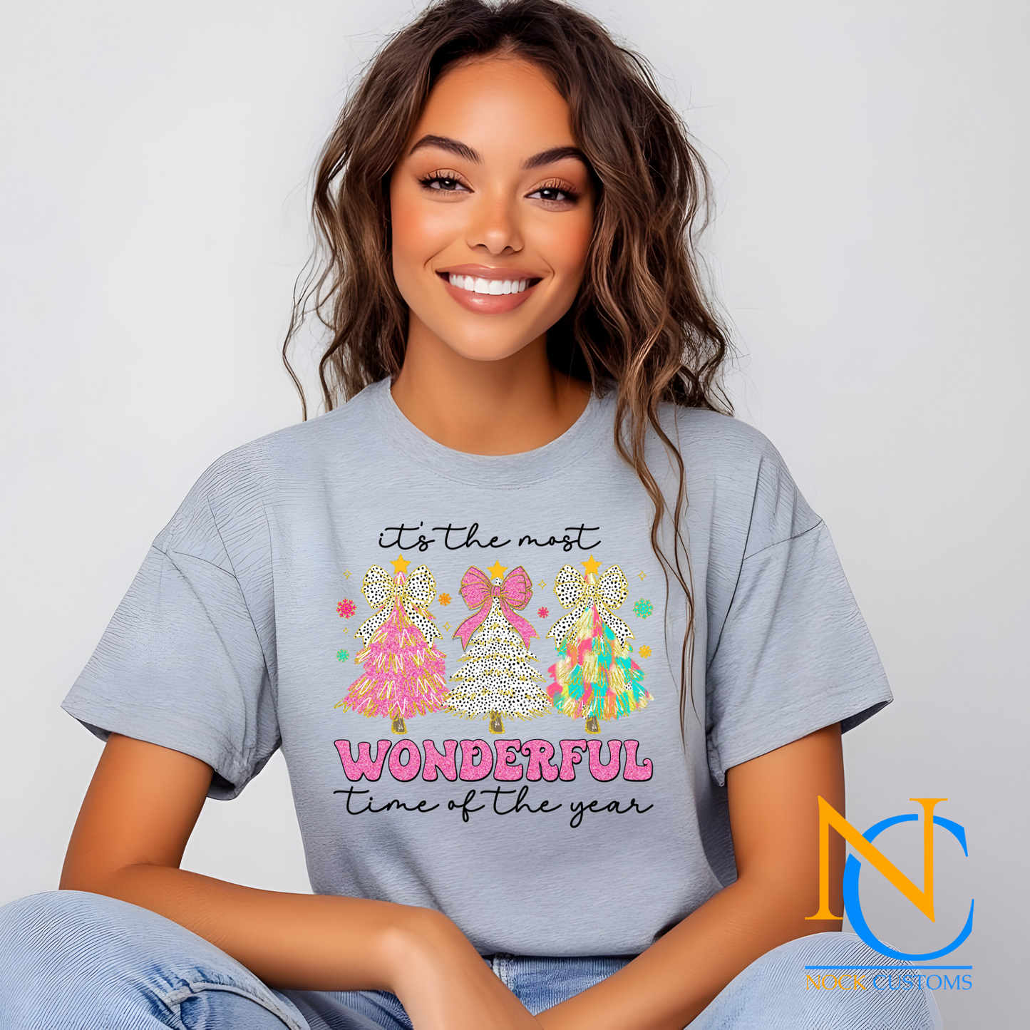 Bright and festive Christmas tree trio design with pink, polka dots, and multicolored trees reading 'It's the most wonderful time of the year' on various t-shirt colors.